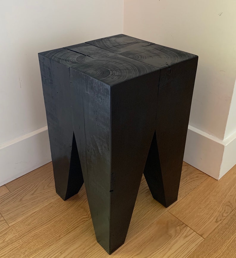May include: Black wooden stool with a square top and four legs. The legs are angled inwards, creating a unique design.