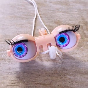 Op de afbeelding: Een paar poppenogen met wimpers, blauwe iris en roze en witte accenten. De ogen zijn bevestigd aan een plastic frame met een wit touwtje.