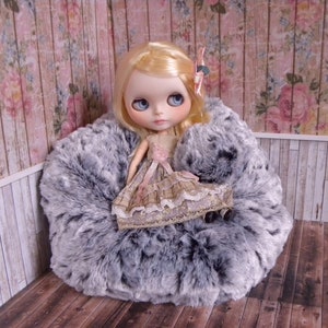 May include: A doll with blonde hair and blue eyes sits in a grey and white furry chair. The doll is wearing a pink and brown dress with lace trim.