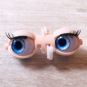 May include: A pair of blue doll eyes with black eyelashes, set in a plastic frame.