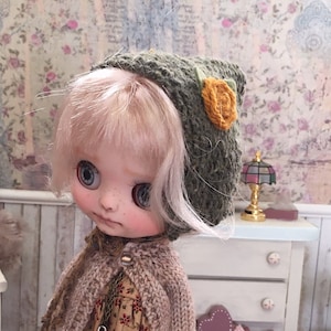 Peut inclure: Une poupée portant un cardigan en tricot marron, une robe à fleurs et un bonnet en tricot vert avec une fleur jaune. La poupée a les cheveux blonds et les yeux bleus. La poupée est debout sur un napperon en crochet blanc.