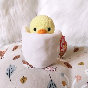 May include: A yellow plush chick toy with black eyes and an orange beak is nestled inside a white plush egg. The egg has a red and white Beanie Baby tag attached to it.
