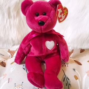May include: A pink Ty Beanie Baby plush bear with a white heart on its chest. The bear is wearing a pink ribbon around its neck and has a Ty tag attached to its ear.