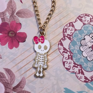 Op de afbeelding: Een gouden kettinghanger met een wit skelet met een roze strik. Het skelet draagt een witte jurk en heeft een roze strik op zijn hoofd.