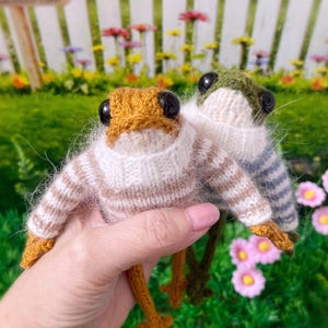 Puede incluir: Dos peluches de rana de punto que llevan suéteres a rayas. La rana de la izquierda lleva un suéter a rayas marrones y blancas y la rana de la derecha lleva un suéter a rayas azules y blancas.