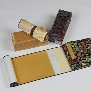 May include: A traditional Chinese calligraphy scroll with a gold background and white paper. The scroll is rolled up and stored in a decorative box with a floral pattern. The box is made of fabric and has a dark blue background with gold accents.
