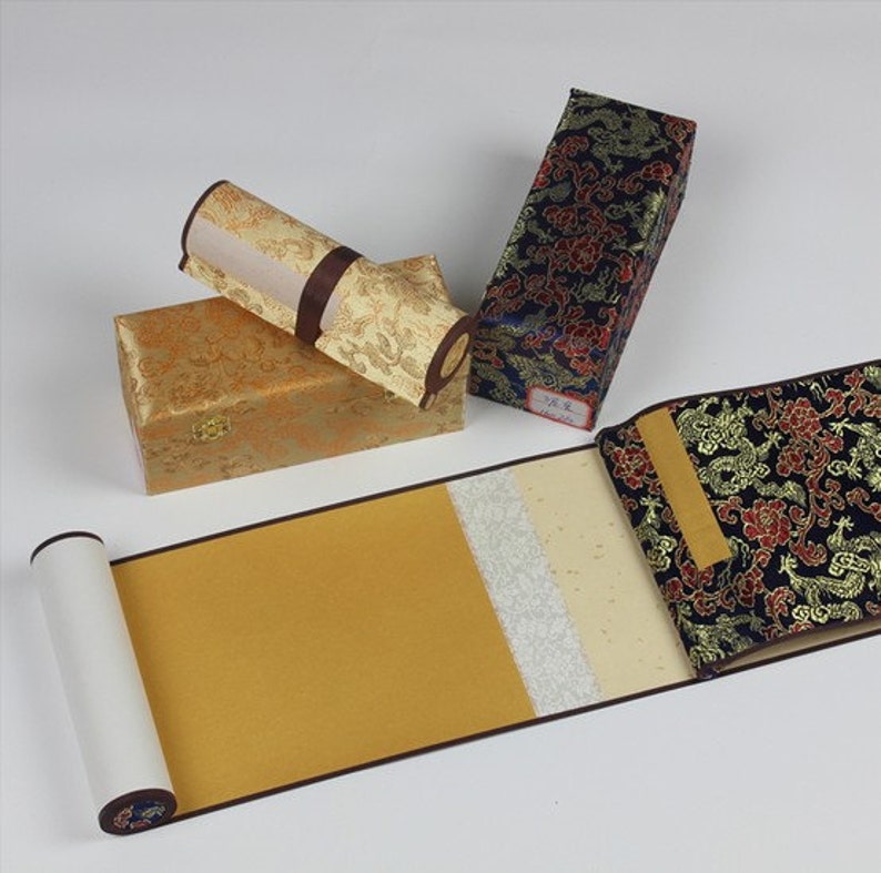 May include: A traditional Chinese calligraphy scroll with a gold background and white paper. The scroll is rolled up and stored in a decorative box with a floral pattern. The box is made of fabric and has a dark blue background with gold accents.