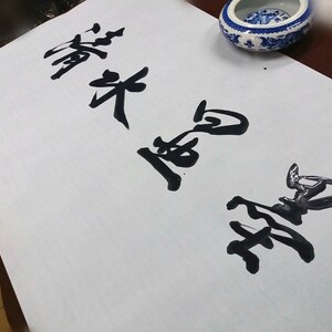 May include: A rectangular sheet of white paper featuring black ink calligraphy characters written in a vertical orientation. The paper is positioned near a small, round, white and blue ceramic bowl with a decorative pattern.