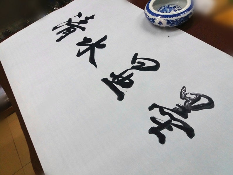 May include: A rectangular sheet of white paper featuring black ink calligraphy characters written in a vertical orientation. The paper is positioned near a small, round, white and blue ceramic bowl with a decorative pattern.