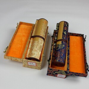 May include: Two decorative boxes with lids open, revealing rolled scrolls inside. The boxes are covered in patterned fabric, one in gold and one in blue with floral designs. The scrolls are wrapped in gold and blue fabric, respectively.