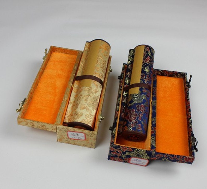 May include: Two decorative boxes with lids open, revealing rolled scrolls inside. The boxes are covered in patterned fabric, one in gold and one in blue with floral designs. The scrolls are wrapped in gold and blue fabric, respectively.