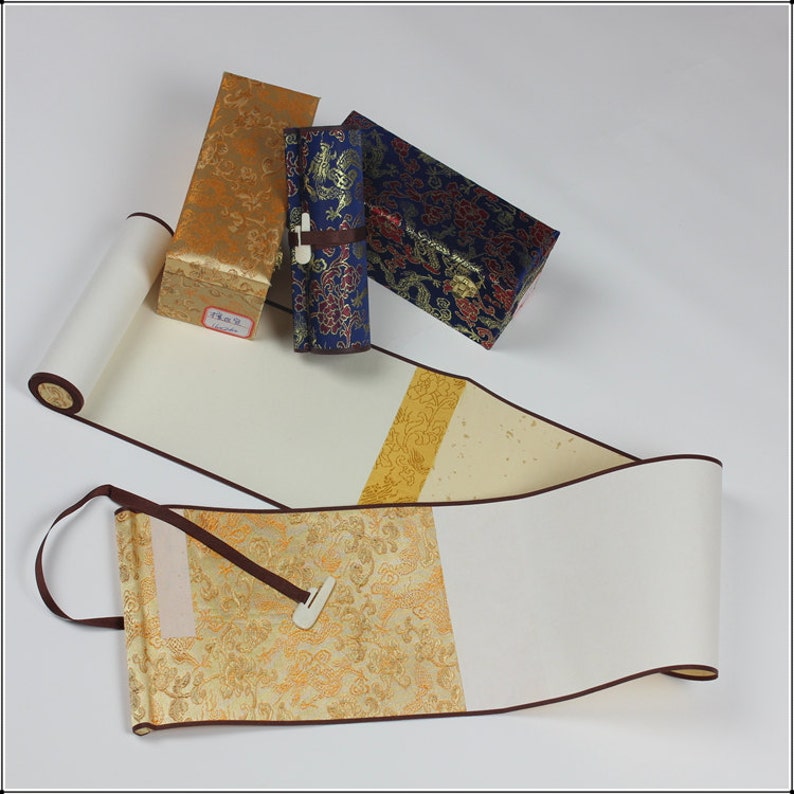 May include: Two white scroll paintings with brown trim and a gold and brown patterned fabric backing. The scrolls are rolled up and tied with a brown ribbon. Three small boxes with gold, blue, and red patterned fabric are also in the image.