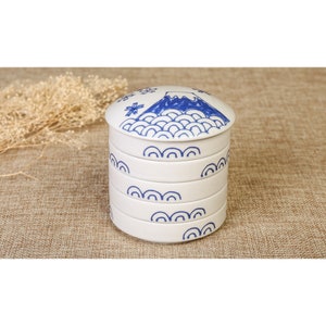 Può includere: Un contenitore di stoccaggio a più livelli in ceramica bianca con un design blu e bianco che presenta una montagna e onde. Il coperchio ha un design blu e bianco di una montagna con un fiocco di neve in cima.
