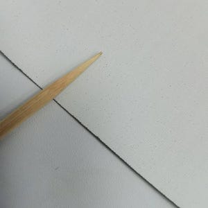 May include: A light gray, textured sheet of material with a fine, speckled pattern across its surface. A thin, pointed wooden tool is positioned diagonally across the sheet, highlighting the texture and the contrast between the wood and the gray.