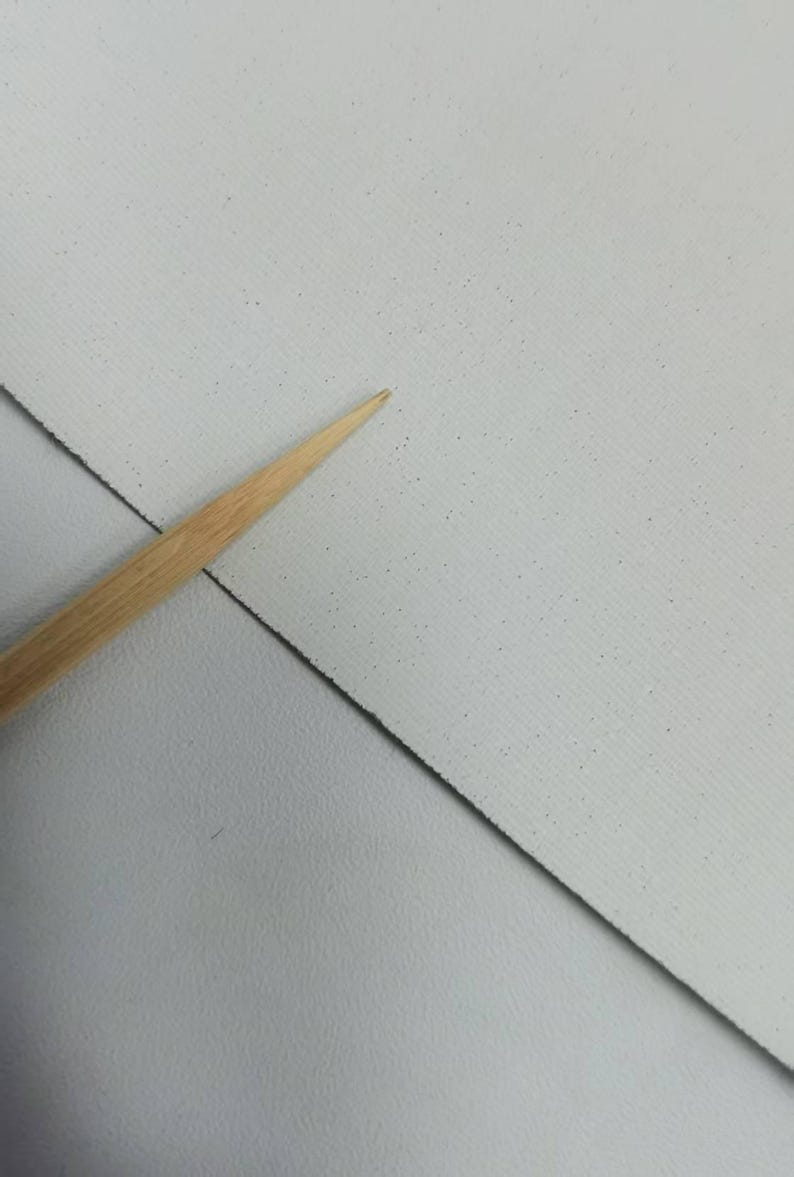 May include: A light gray, textured sheet of material with a fine, speckled pattern across its surface. A thin, pointed wooden tool is positioned diagonally across the sheet, highlighting the texture and the contrast between the wood and the gray.