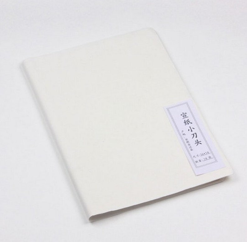 May include: A white, blank, paper notebook with a small, rectangular label on the front. The label is white with black text that reads "宣紙 小刀 光" and has additional text in Japanese.