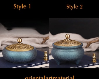 May include: Two styles of blue and gold incense burners. The incense burner on the left has a round base and a lid with a floral design. The incense burner on the right has a round base with three legs and a lid with a floral design. Both incense burners have a small incense stick holder.