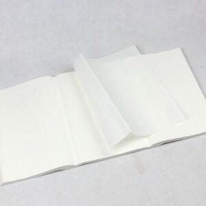 May include: An open notebook with a soft, off-white cover and several pages turned. The pages are bright white, and the notebook lies flat on a light grey surface. The notebook appears to be unused.