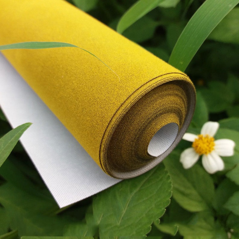 May include: A roll of mustard yellow fabric with a textured surface and a smooth white backing. The material is tightly wound into a cylindrical shape, displaying a consistent golden-yellow hue across the outer layer and a clean white interior.