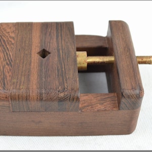 May include: A wooden clamp with a brass screw and handle. The clamp is made of dark wood and has a square shape.