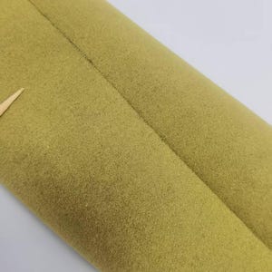 May include: A roll of olive green fabric with a soft, felt-like texture. The material features a visible seam running vertically along the length of the roll, with a small, light-colored pointed object protruding from the left side of the fabric.