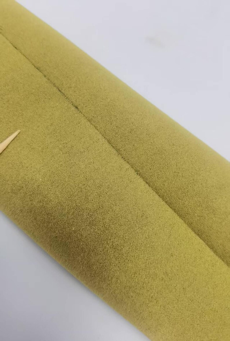 May include: A roll of olive green fabric with a soft, felt-like texture. The material features a visible seam running vertically along the length of the roll, with a small, light-colored pointed object protruding from the left side of the fabric.