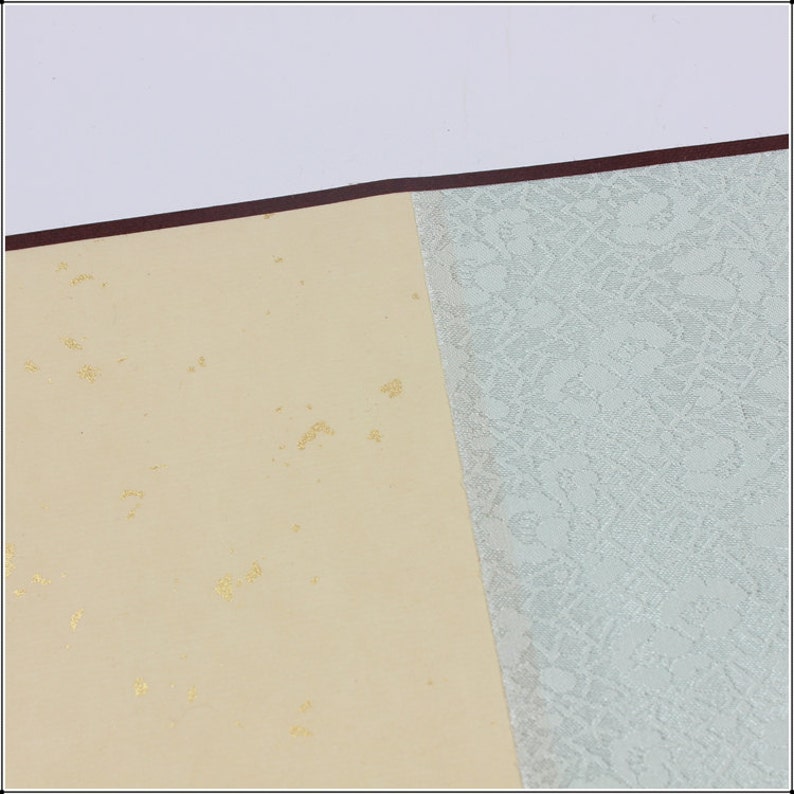 May include: A close-up of a cream-colored paper with gold flecks. The paper is attached to a light blue paper with a textured pattern.