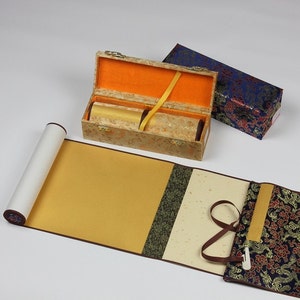 May include: A traditional Chinese scroll painting set with a gold and brown scroll, a gold and brown storage box, and a blue and gold storage box. The scroll is rolled up and tied with a brown ribbon.