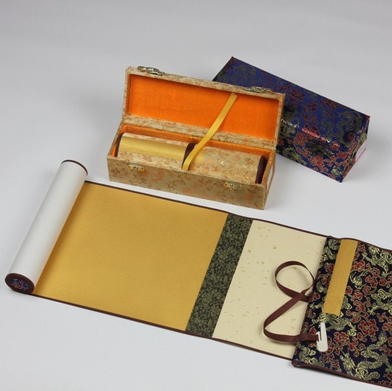 May include: A traditional Chinese scroll painting set with a gold and brown scroll, a gold and brown storage box, and a blue and gold storage box. The scroll is rolled up and tied with a brown ribbon.