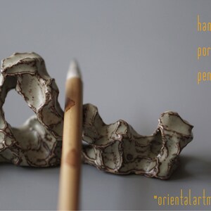 Op de afbeelding: Een handgemaakte porseleinen penhouder met een unieke, organische vorm. De penhouder is gebroken wit met bruine accenten en een textuur. Een houten pen is ervoor geplaatst. Tekst op de afbeelding luidt "handmade porcelain pen holder" en "orientalartmaterial".