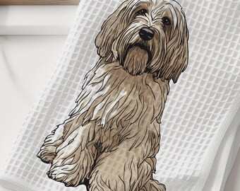 May include: White waffle-weave kitchen towel featuring a detailed illustration of a tan-colored dog. The dog is depicted in a seated position with long, flowing fur. The towel is folded and placed on a white surface, with a wooden cutting board and a white vase with greenery in the background.