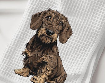 May include: A white waffle-weave kitchen towel featuring a detailed illustration of a brown wire-haired dachshund sitting upright. The dog has a shaggy coat, long floppy ears, and a dark nose, centered on the textured fabric of the towel.