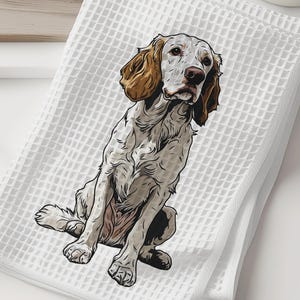 May include: White waffle-weave kitchen towel featuring a detailed illustration of a dog. The dog has brown and white fur, with floppy ears and a focused expression. The towel is folded and sits on a white surface, suggesting a home decor or pet-themed product.