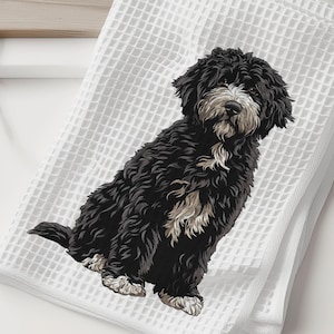 May include: White waffle-weave towel featuring a detailed illustration of a black and white dog. The dog is sitting, with its fluffy fur and expressive face prominently displayed. The towel is folded and placed on a white surface, with a wooden cutting board and a white vase with greenery in the background.