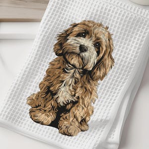 May include: White waffle-weave towel featuring a detailed illustration of a brown and white Cavapoo puppy. The puppy is sitting and looking towards the viewer. The towel is folded and placed on a white surface.