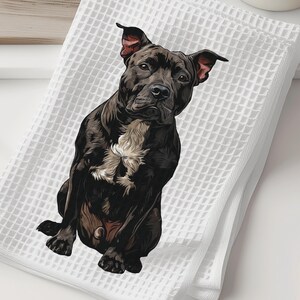 May include: White waffle-weave towel featuring a detailed illustration of a black and brown pit bull. The dog has a white patch on its chest and is sitting upright. The towel is folded and placed on a white surface.