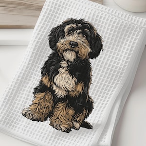 May include: White waffle-weave towel featuring a detailed illustration of a seated dog with black, brown, and tan fur. The dog is centered on the folded towel, which is placed on a white surface. A wooden cutting board and a white vase with greenery are in the background.