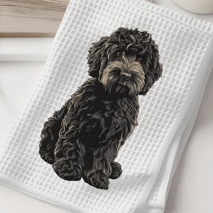 May include: White waffle-weave towel featuring a detailed illustration of a black dog with a tan muzzle. The dog is sitting, facing forward. The towel is folded and stacked, with a wooden cutting board and a white vase with greenery in the background.