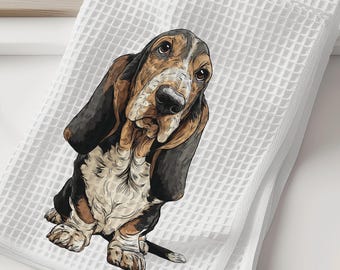 May include: A white waffle-weave kitchen towel featuring a detailed, colorful illustration of a Basset Hound dog. The dog has long, drooping ears, a black, brown, and white coat, and is depicted in a seated position looking upward.