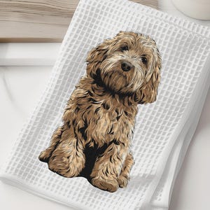 May include: White waffle-weave towel featuring a detailed illustration of a brown Cockapoo dog. The dog is sitting upright with a focused expression. The towel is folded and placed on a white surface, with a wooden cutting board and a white vase with greenery in the background.