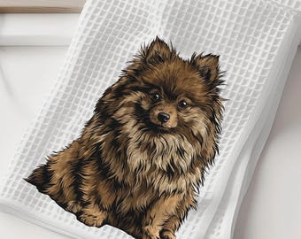 May include: White waffle-weave towel featuring a detailed illustration of a brown Pomeranian dog. The dog is depicted in a sitting position with fluffy fur. The towel is folded and placed on a white surface, suggesting a bathroom or kitchen setting.