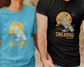 May include: A couple wearing matching t-shirts with a graphic of a raven and the text "Swearing Helps". The woman is wearing a light blue t-shirt and the man is wearing a black t-shirt. They are walking on a dirt path in a forest.