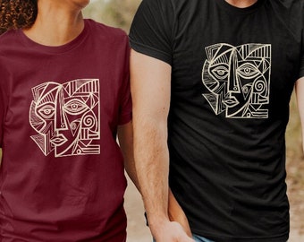 May include: A couple wearing matching t-shirts with a geometric abstract face design in white on a black and maroon background. The man is wearing a black t-shirt and the woman is wearing a maroon t-shirt. They are walking on a dirt path in a forest.
