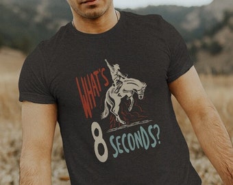 May include: A dark grey t-shirt with a vintage-style graphic of a cowboy riding a bucking bronco. The graphic includes the text "What's 8 Seconds?"