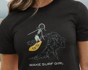 May include: Black t-shirt featuring a stick figure surfing on a yellow surfboard, with the text "WAKE SURF GIRL" below the graphic. The shirt is worn by a person in a natural outdoor setting.