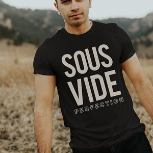 May include: A man wearing a black t-shirt with the text "Sous Vide Perfection" in white. He is standing in a field of dry grass.