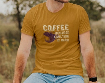 May include: A man wearing a mustard yellow t-shirt with the text "COFFEE because adulting is hard" and a graphic of a coffee cup. He is sitting on a wooden fence in a field.