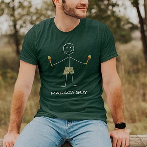 May include: A green t-shirt with a stick figure holding maracas and the text "MARACA GUY". The person wearing the shirt is sitting on a wooden fence.