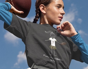 May include: A young person wearing a black t-shirt with a stick figure illustration of a girl playing flag football. The shirt says "Flag Football Girl". The person is holding a brown football and wearing white shorts with a blue belt.