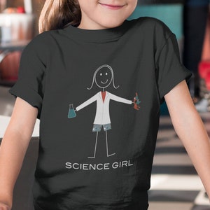 Puede incluir: Una persona joven lleva una camiseta negra con una ilustraci&oacute;n de un cient&iacute;fico de palo blanco sosteniendo un vaso de precipitados y un tubo de ensayo. La camiseta tiene el texto "SCIENCE GIRL" impreso en la parte inferior.